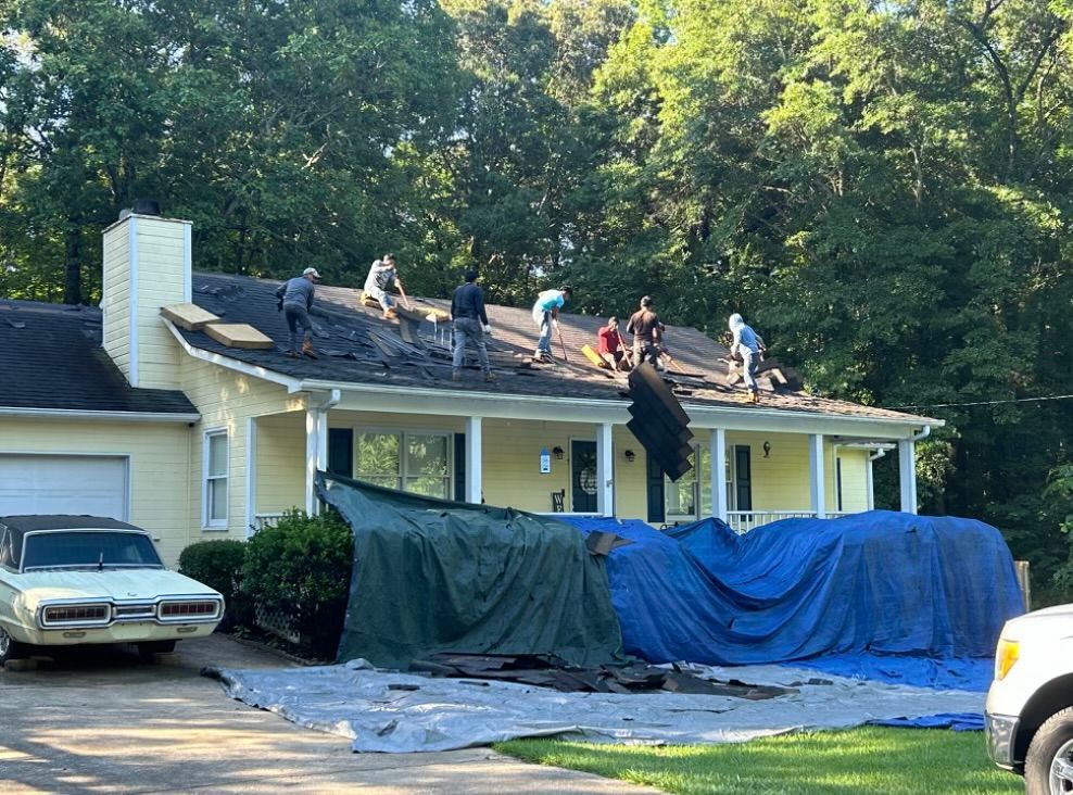 Roofers working on a yellow house. Tarps cover the lawn; a vintage car sits in the driveway.