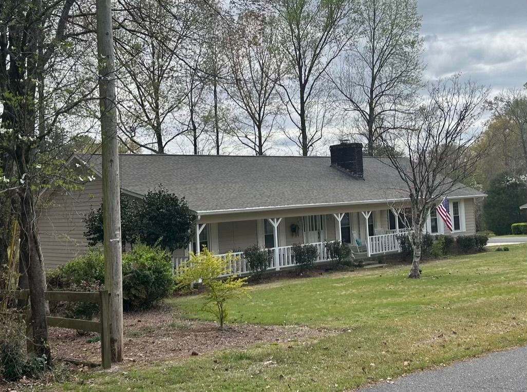 A beige ranch-style house with a long front porch, chimney, and an American flag.