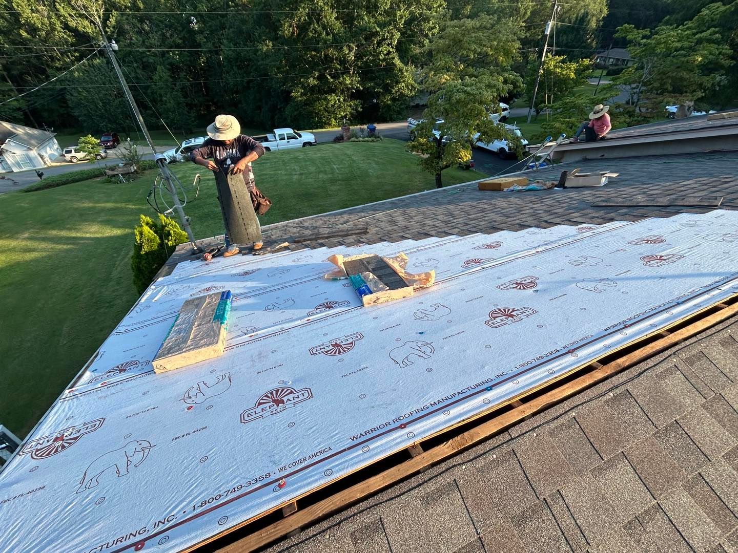Roofers on a rooftop, laying down underlayment. Sunny day, green yard in the background.