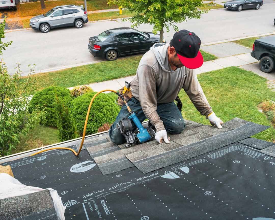 Roofer kneeling, installing shingles on a house roof with a pneumatic nailer.