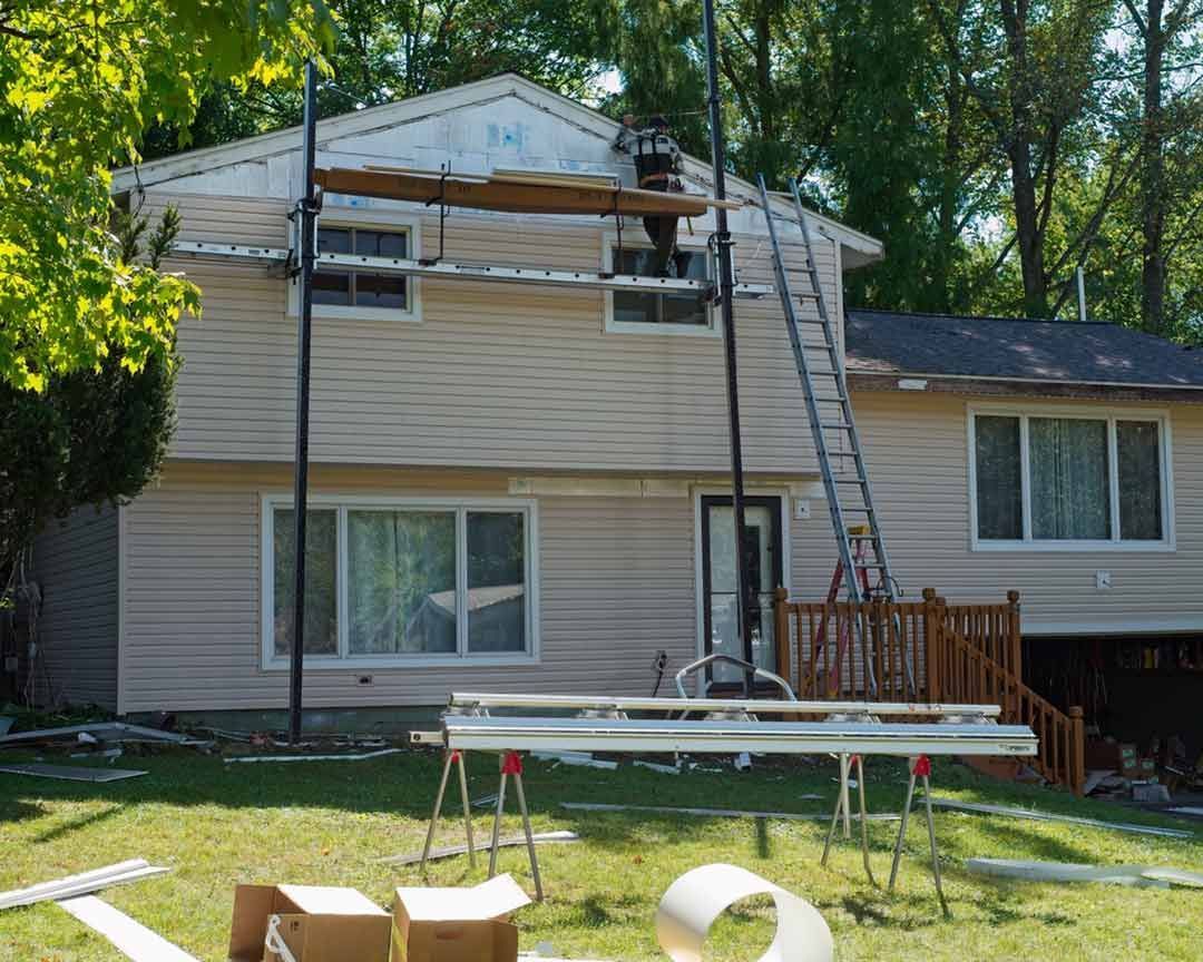 House under construction; siding being replaced. Scaffolding, ladder, tools visible. Sunny day.