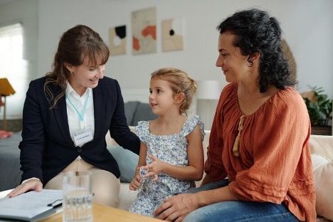 Woman talking with a child while another woman smiles on a sofa in a bright living room.