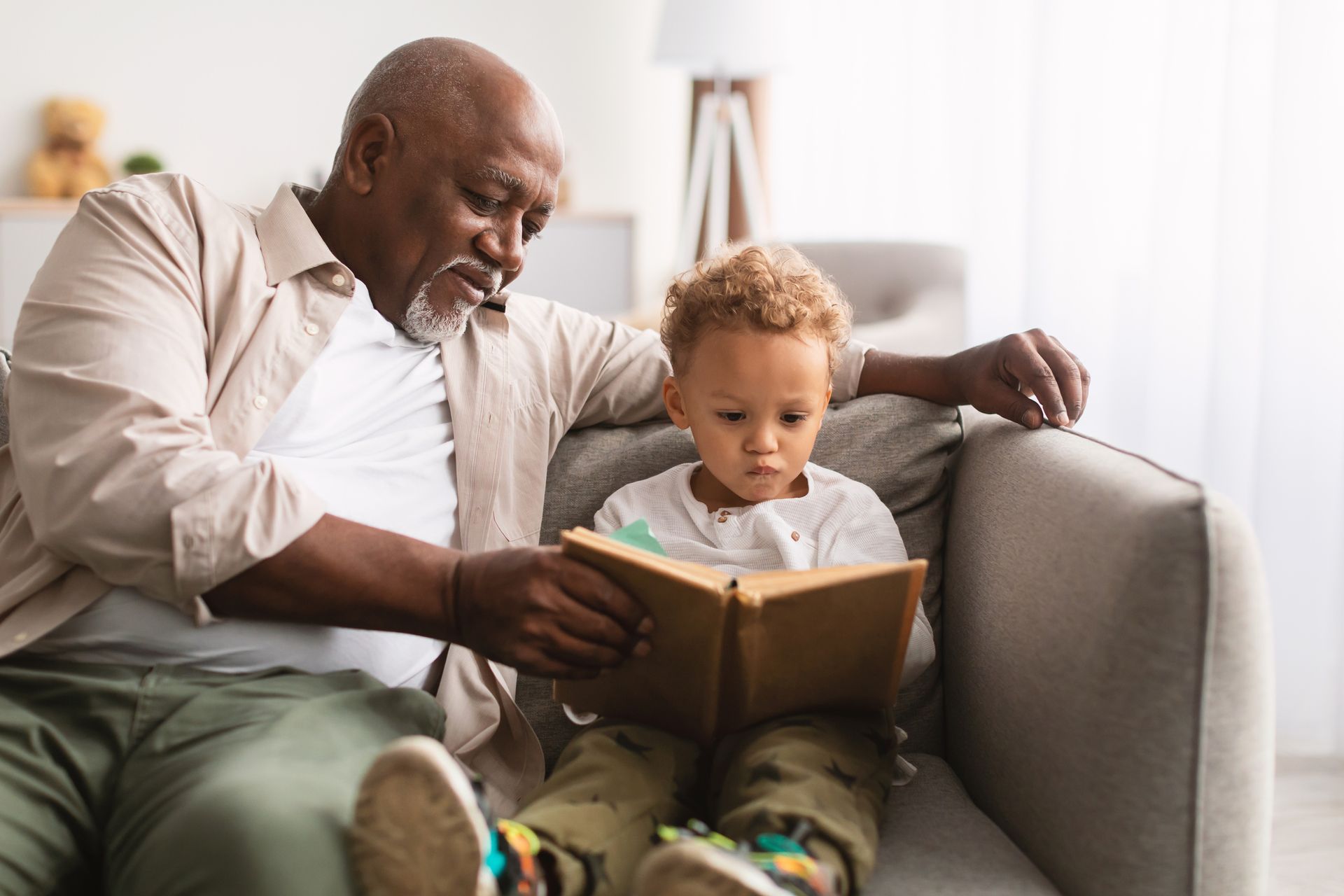 An elderly man is reading a book to a young boy while sitting on a couch.
