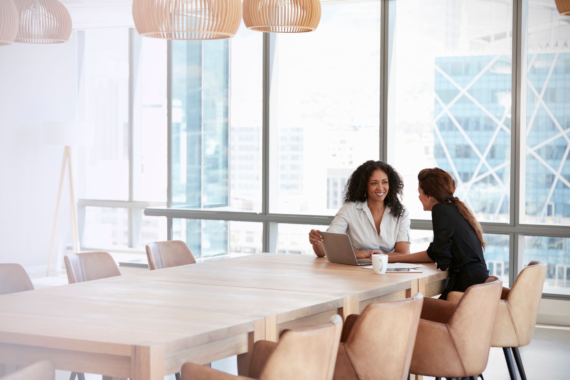 Two women are sitting at a long table in a conference room talking to each other.