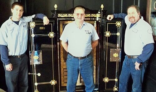 Three men in work uniforms stand in front of large black safes.