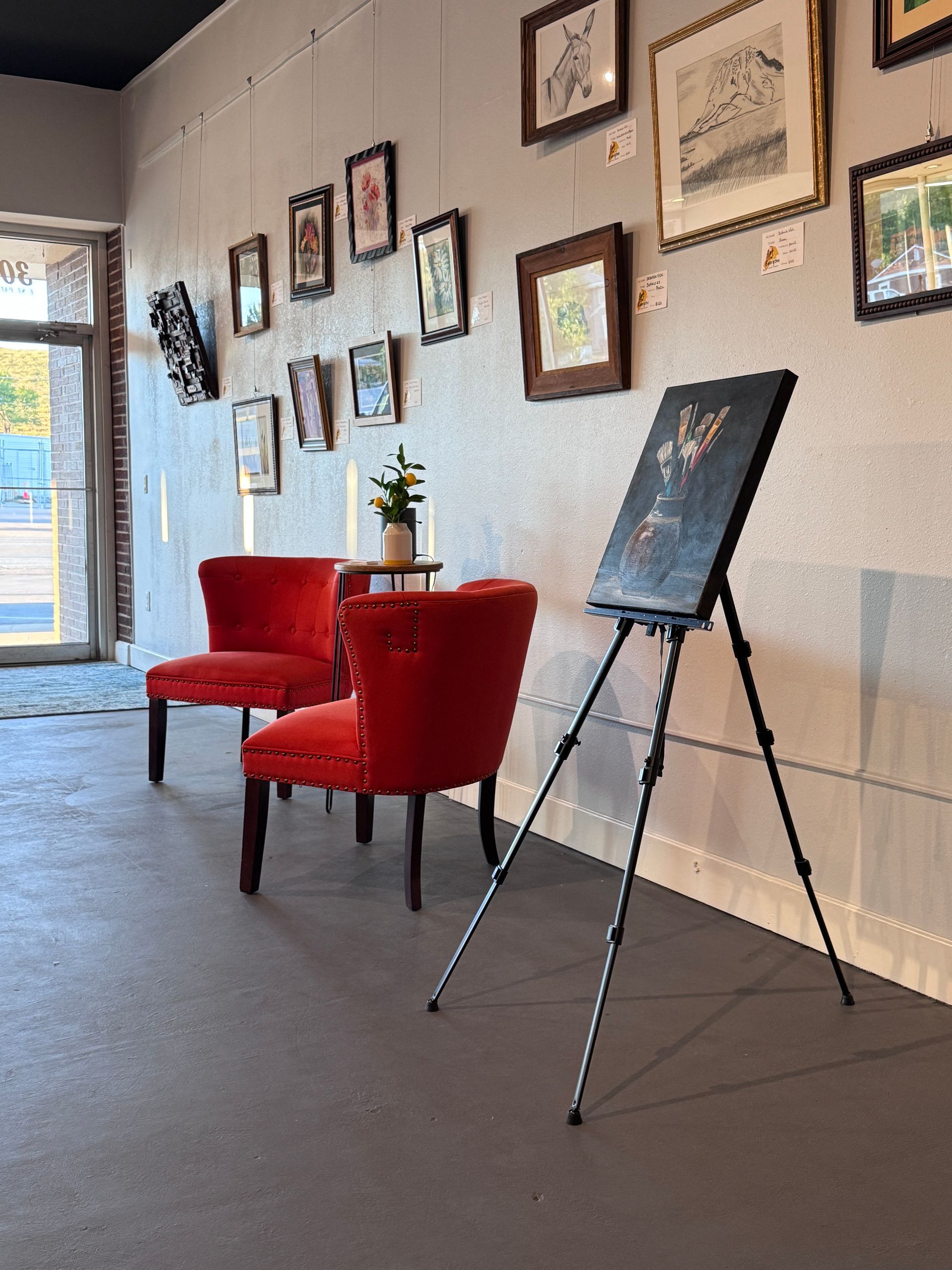Art gallery interior: red chairs, artwork on walls, easel holding a painting, and a small plant.
