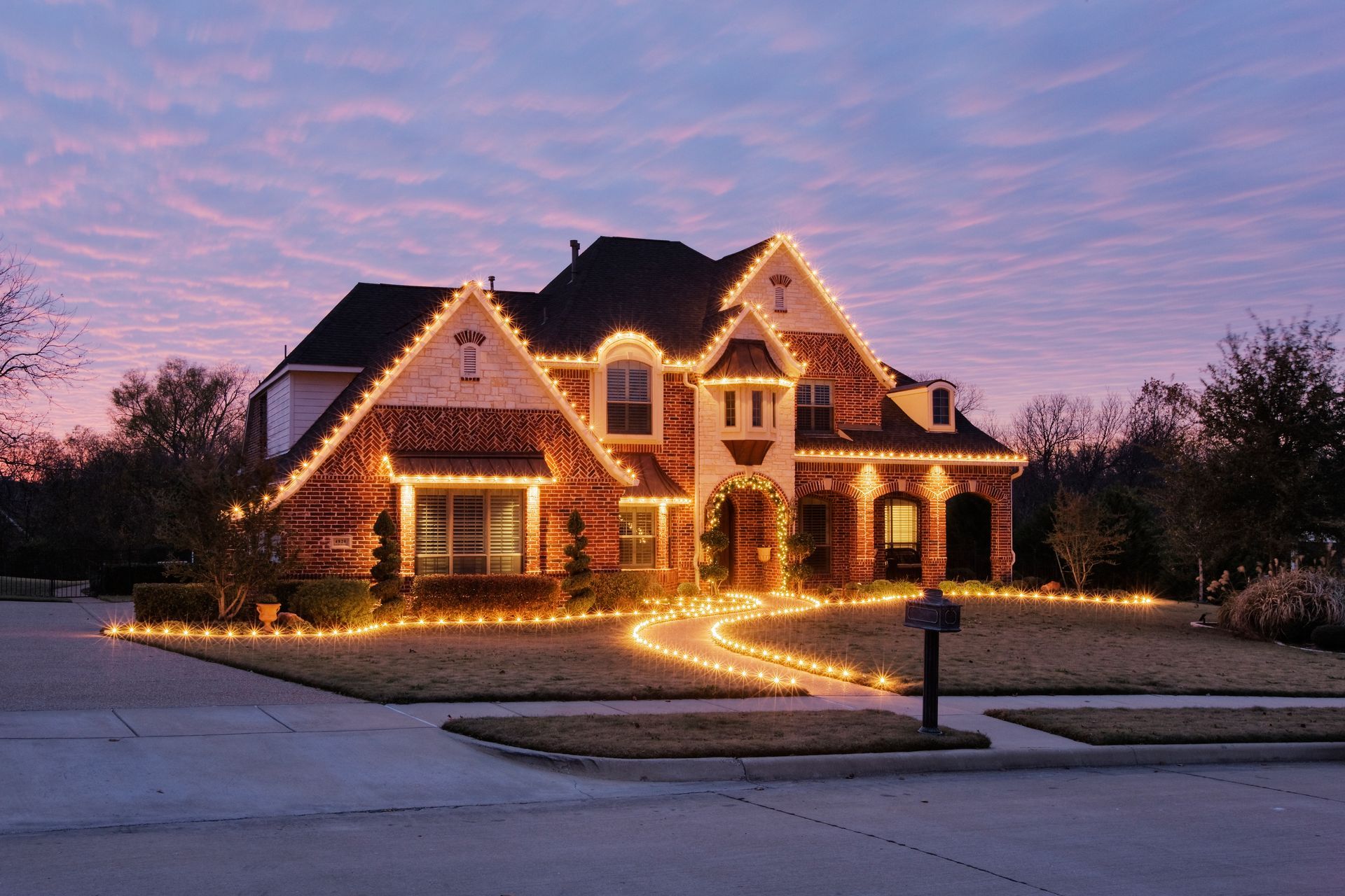 House decorated with Christmas lights at dusk, lit walkway, suburban setting.
