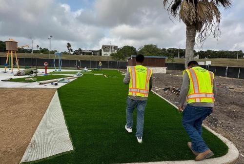 Two men in yellow vests are walking on artificial grass