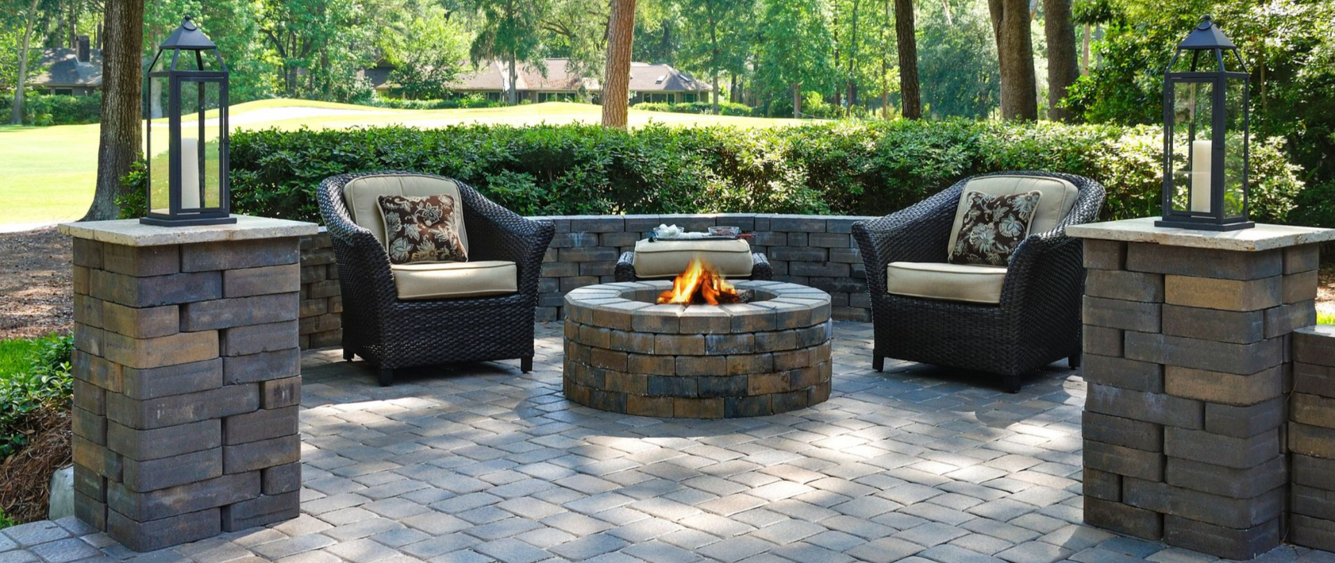 Patio with fire pit, chairs, lanterns, and stone pillars, in a backyard setting.