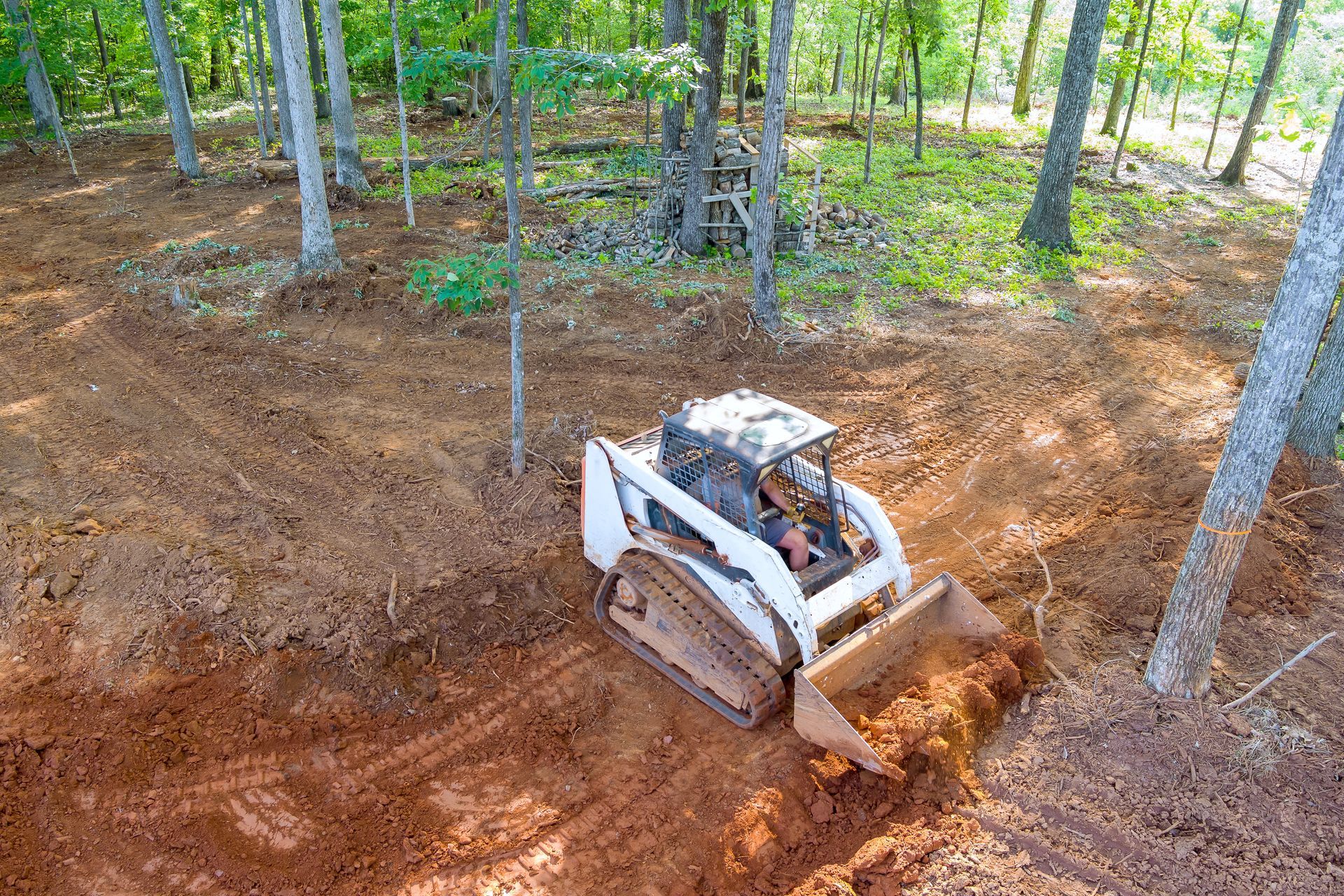 White skid-steer loader digging dirt in a wooded area, with trees and a brown earth mound nearby