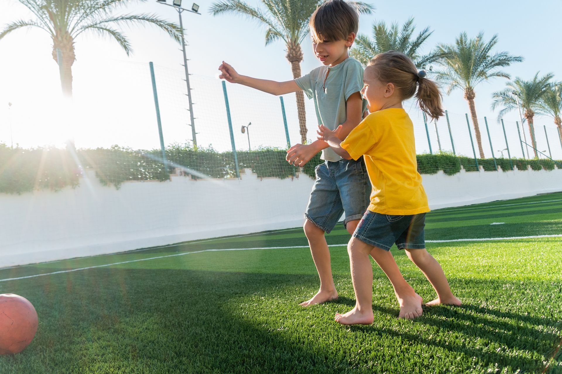Two children play with a red ball on a bright, sunny turf field lined with palm trees.