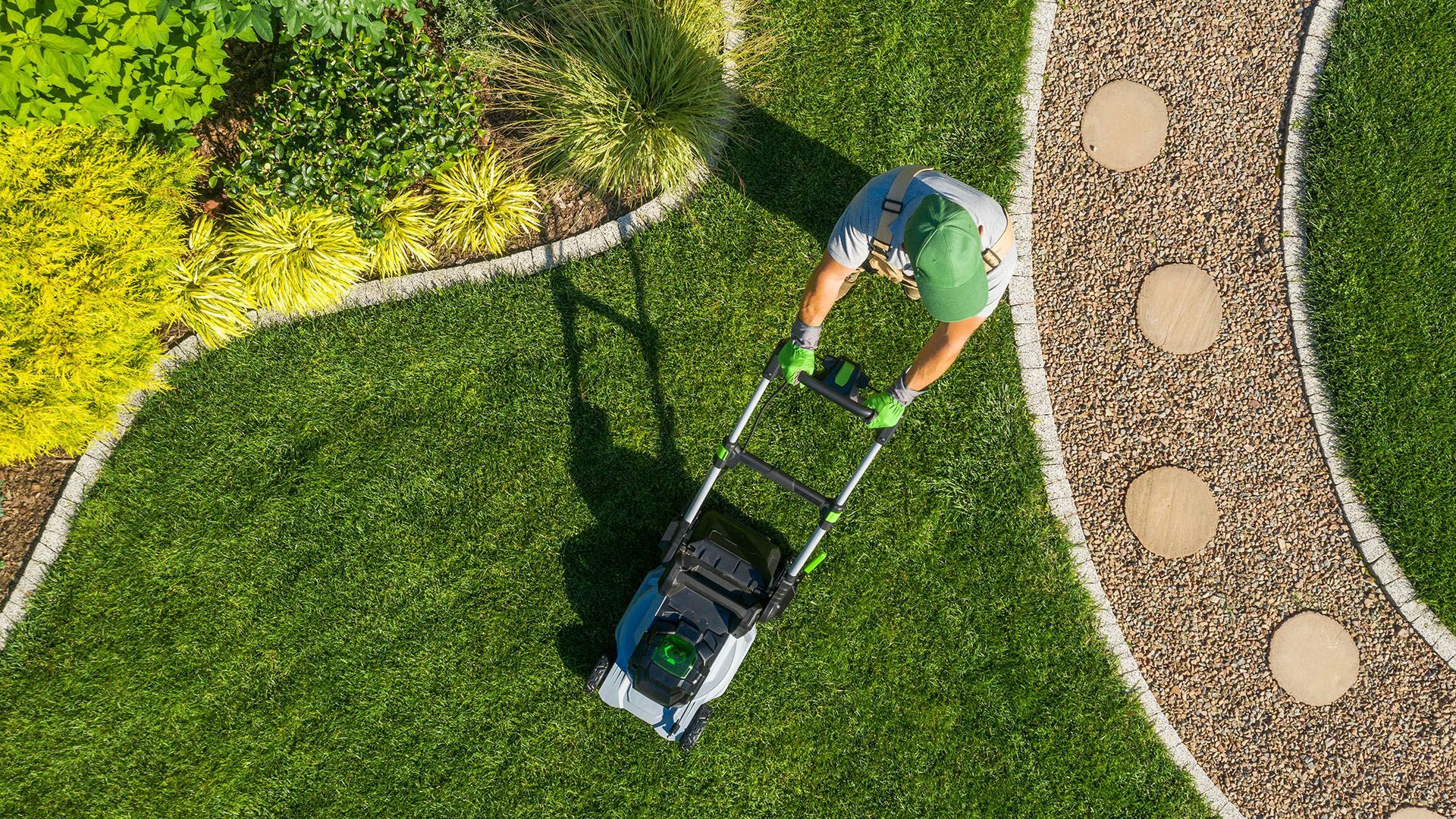 Person mowing a vibrant green lawn with an electric mower near a flower bed and stone path.