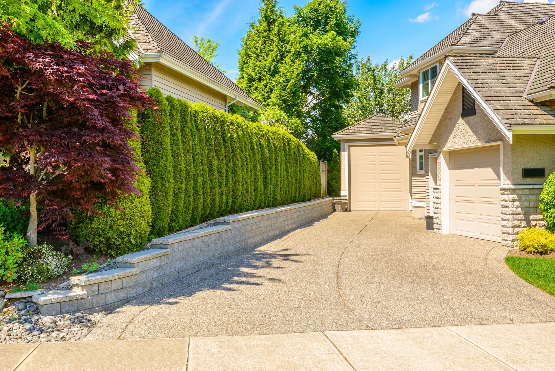 Residential driveway beside a beige house and garage, lined with green hedges and trees