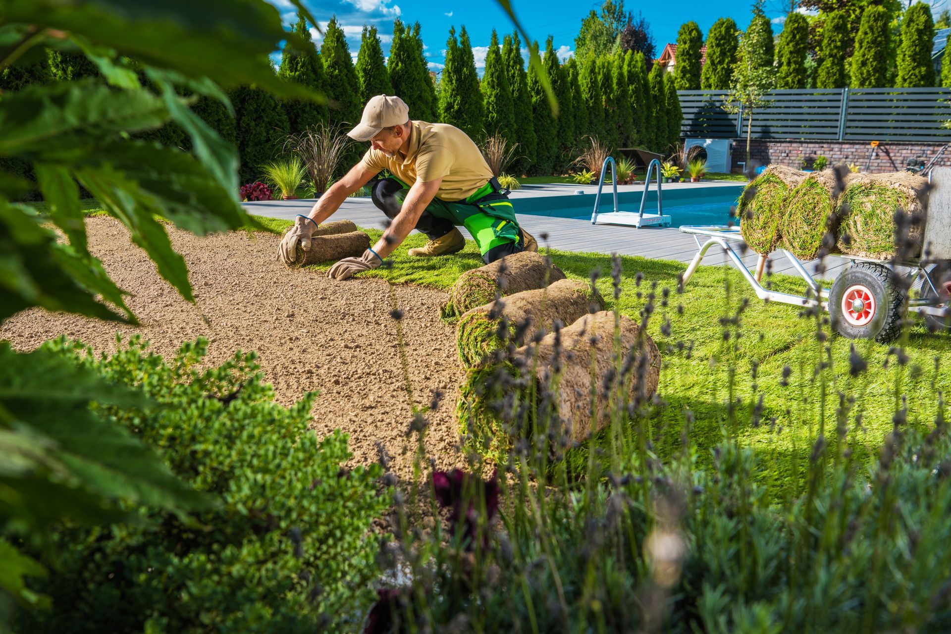 Man laying sod in a backyard near a pool. He's wearing a hat, green pants, and a yellow shirt.