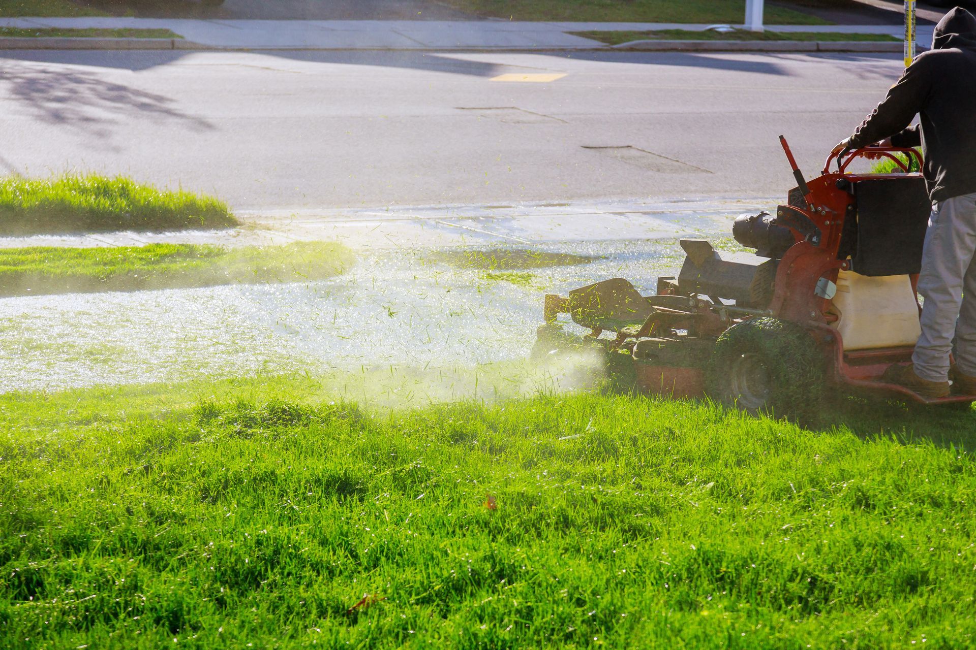 Person mowing grass near a street, with a riding mower, leaving a trail of freshly cut grass.