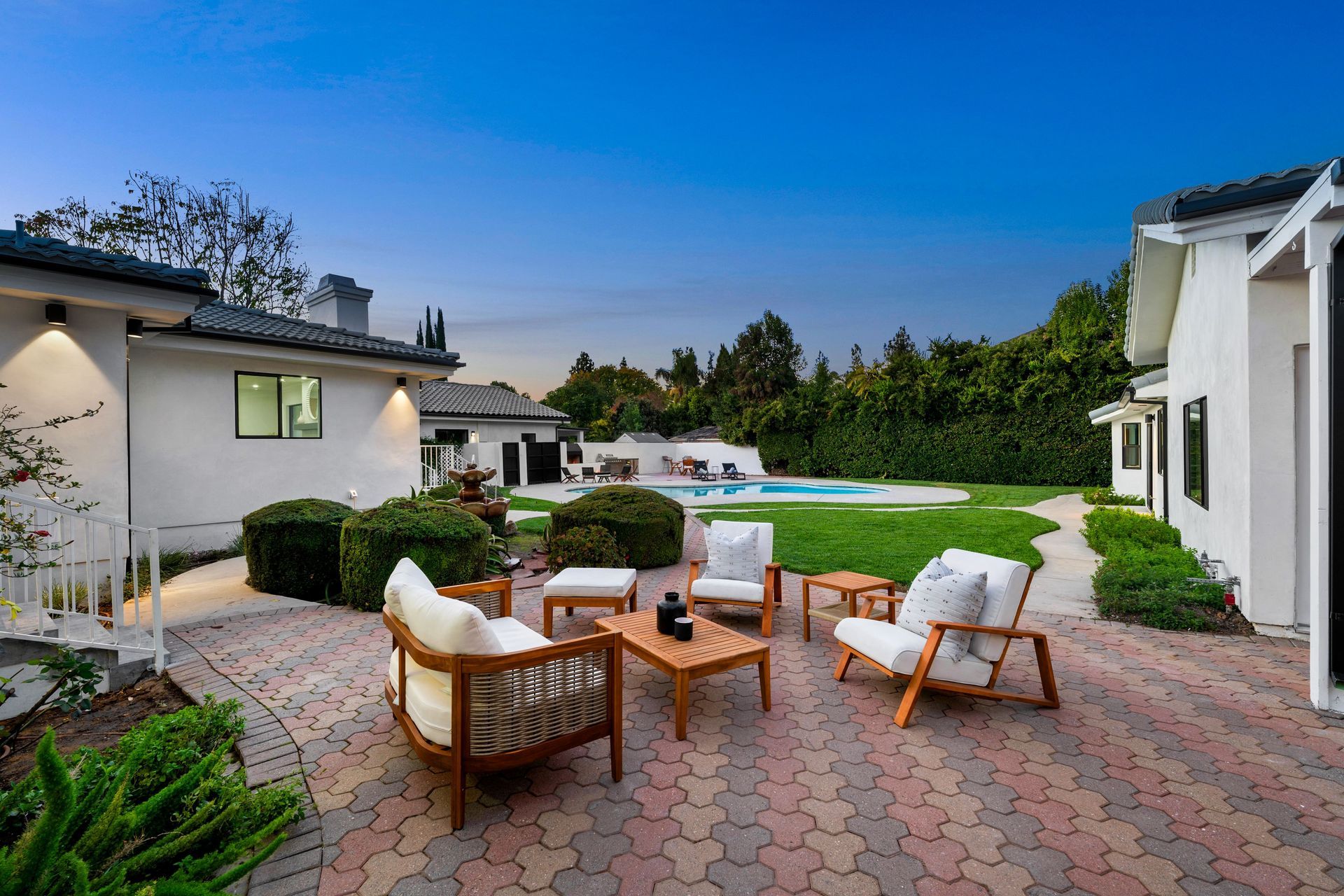 Patio with wicker chairs and fire pit beside a lawn at dusk, framed by modern white houses