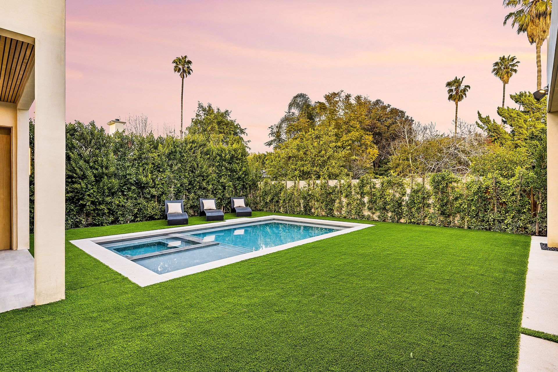 Backyard with a rectangular pool, lounge chairs, lush green lawn, and palm trees at sunset