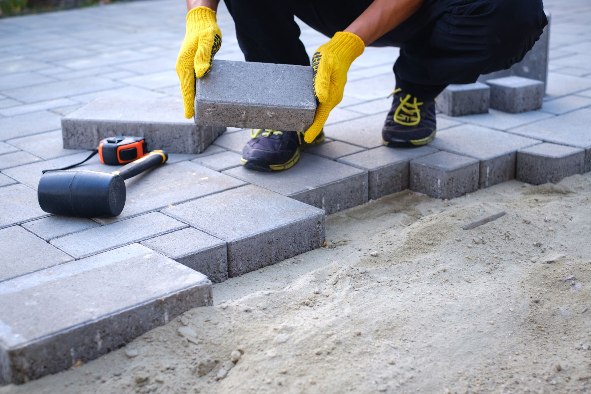 Person placing a paving stone, yellow gloves, black pants, sandy ground.