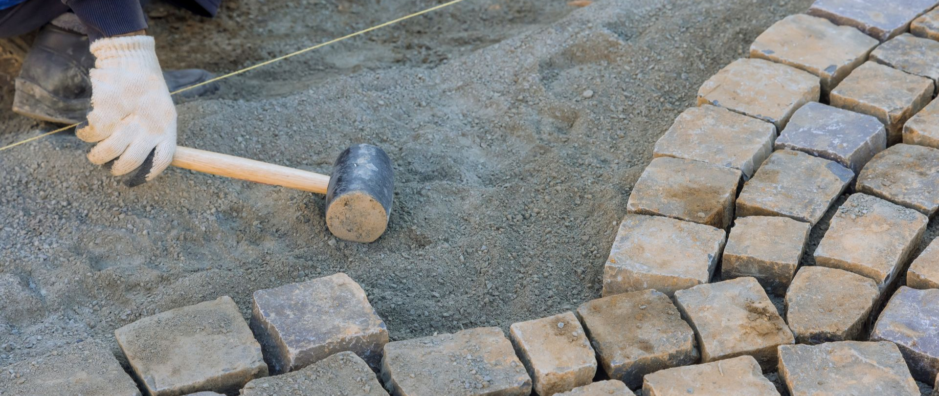 Person laying cobblestones with a mallet on a gravel surface; curved pathway being built.