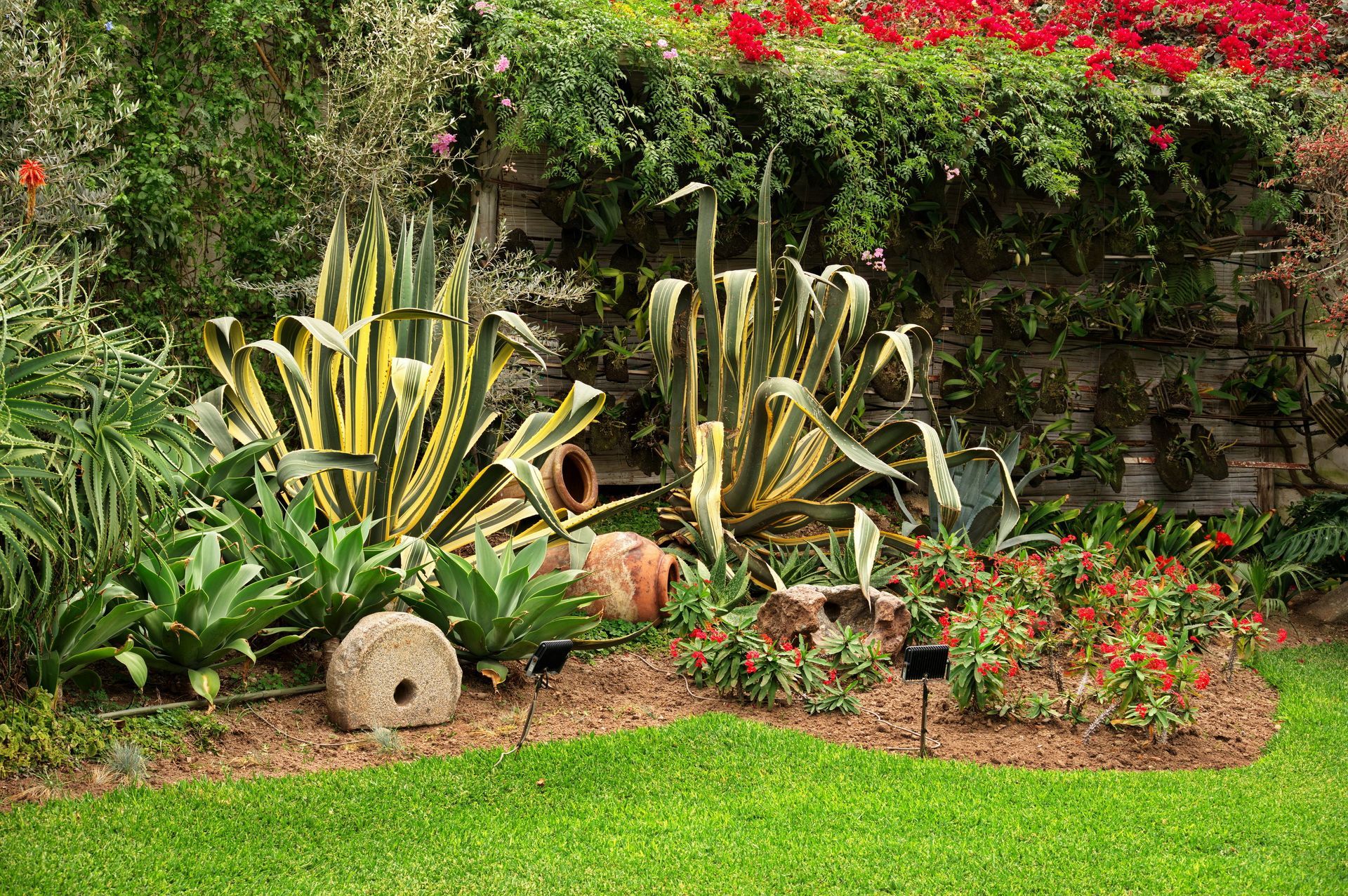 Lush garden bed with green and yellow plants, red flowers, stone edging, and a lawn in front