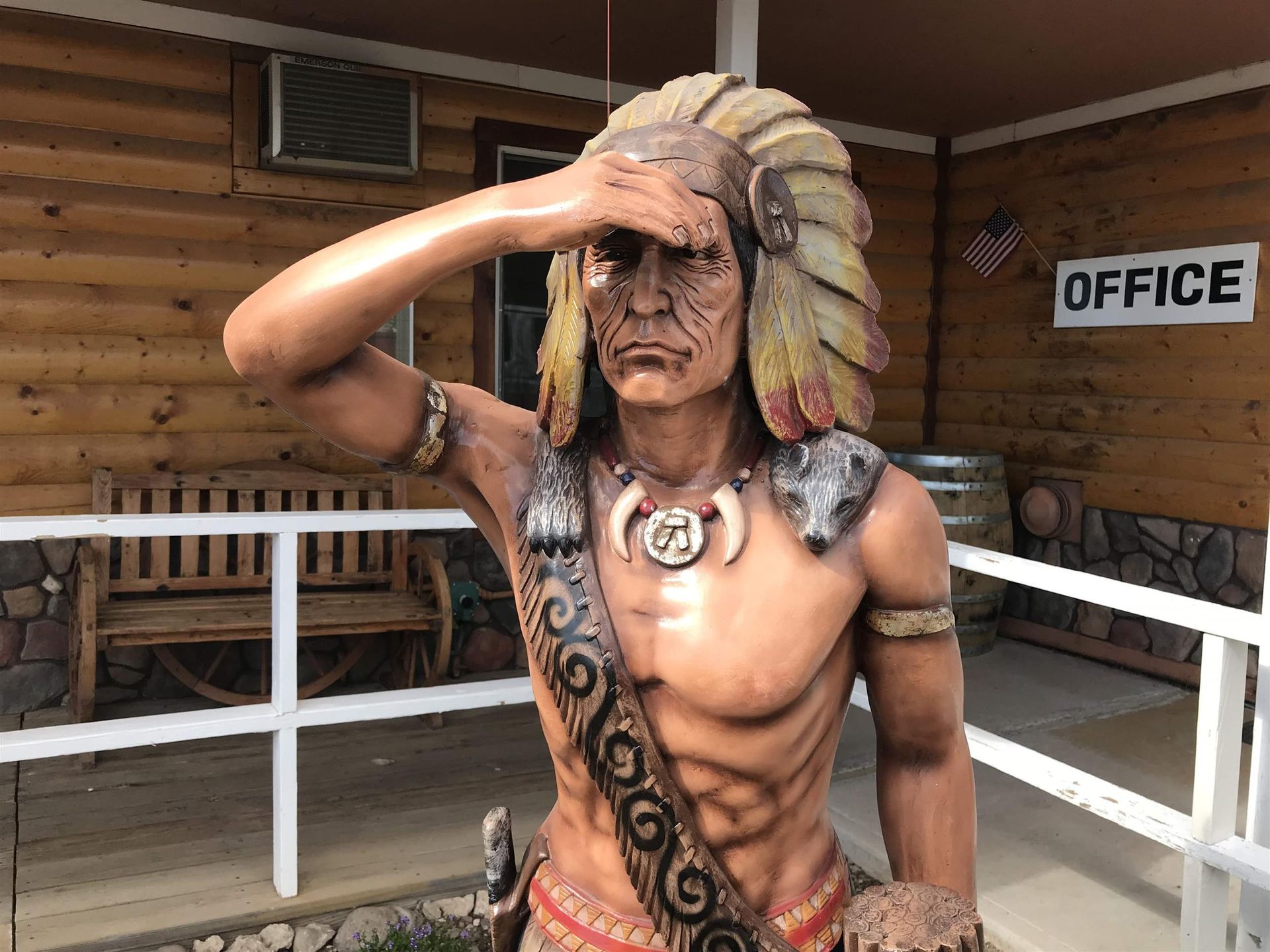 A statue of a native american is saluting in front of a wooden building.
