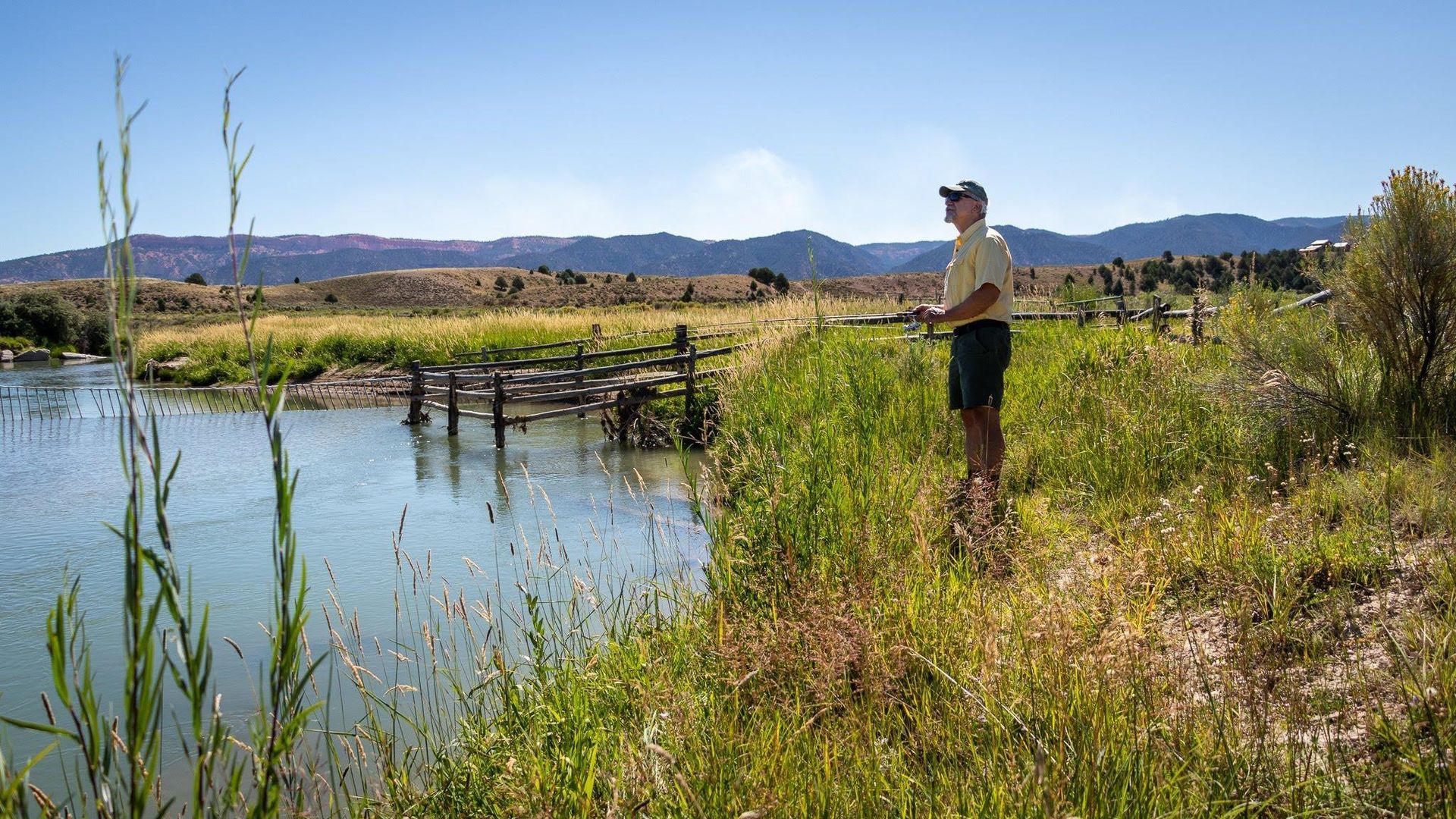 A man is fishing in a pond with mountains in the background.