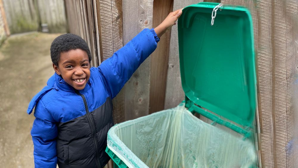 A young boy in a blue jacket is standing next to a green trash can.