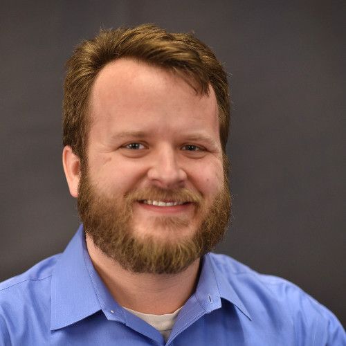 A man with a beard wearing a blue shirt is smiling for the camera.