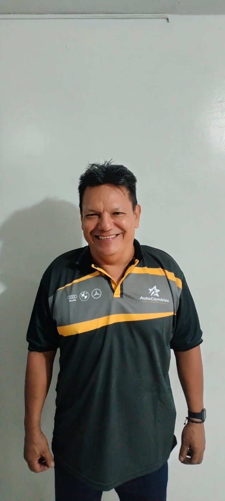 A man wearing a black and yellow polo shirt is standing in front of a white wall.