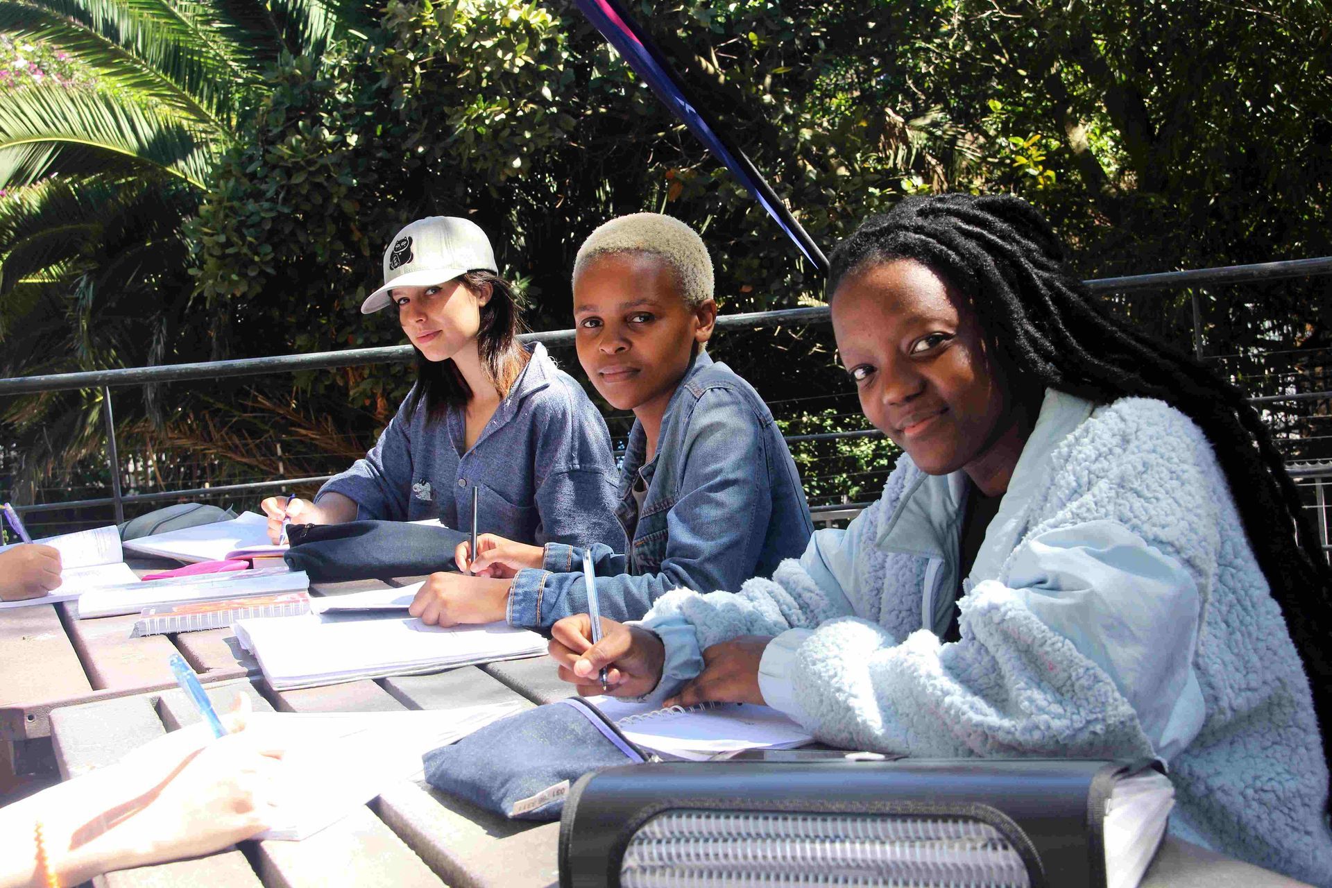 Three people sit at an outdoor table, writing on papers in a garden setting.
