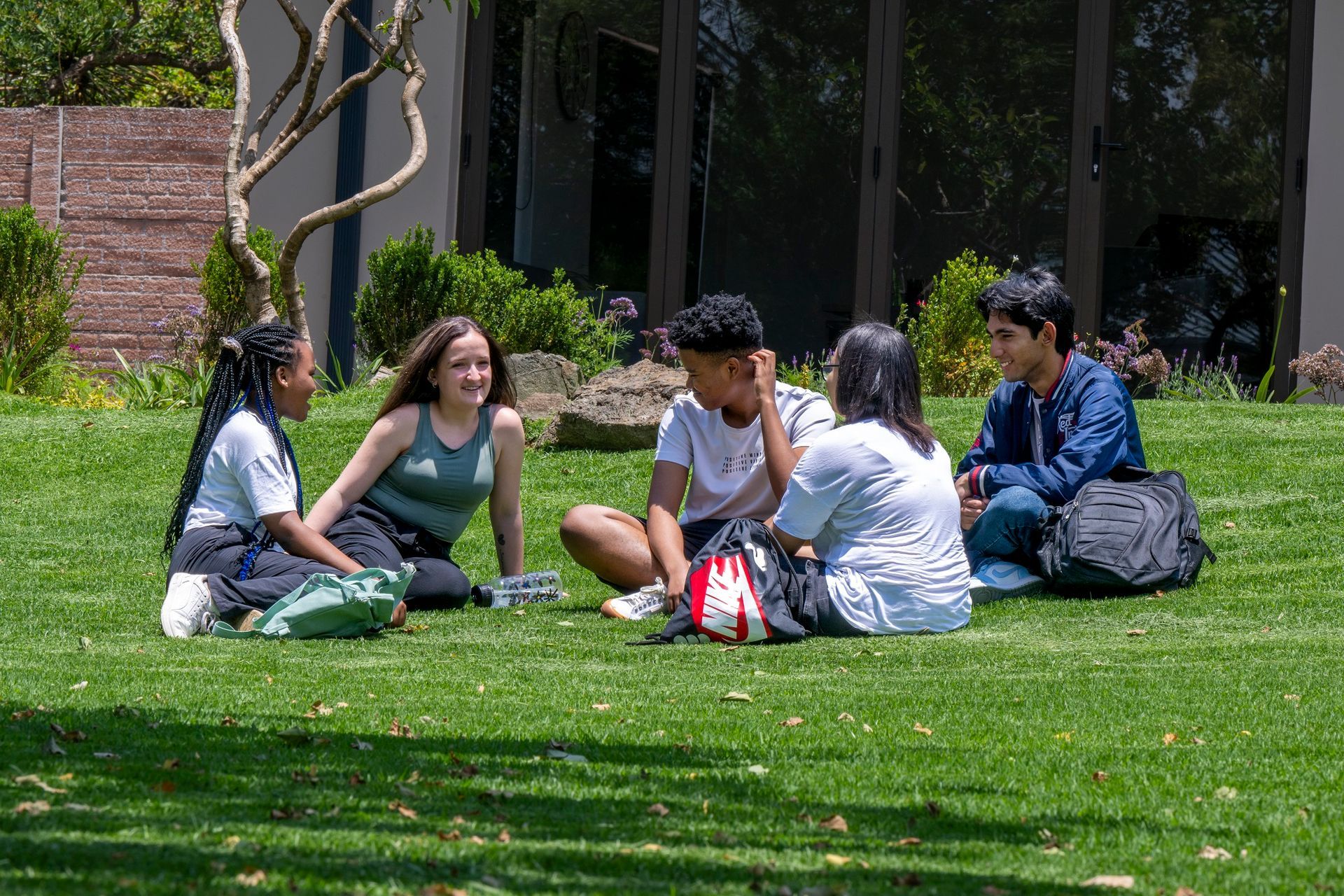 Five students sit on grass, talking. Building in background. Sunny day.
