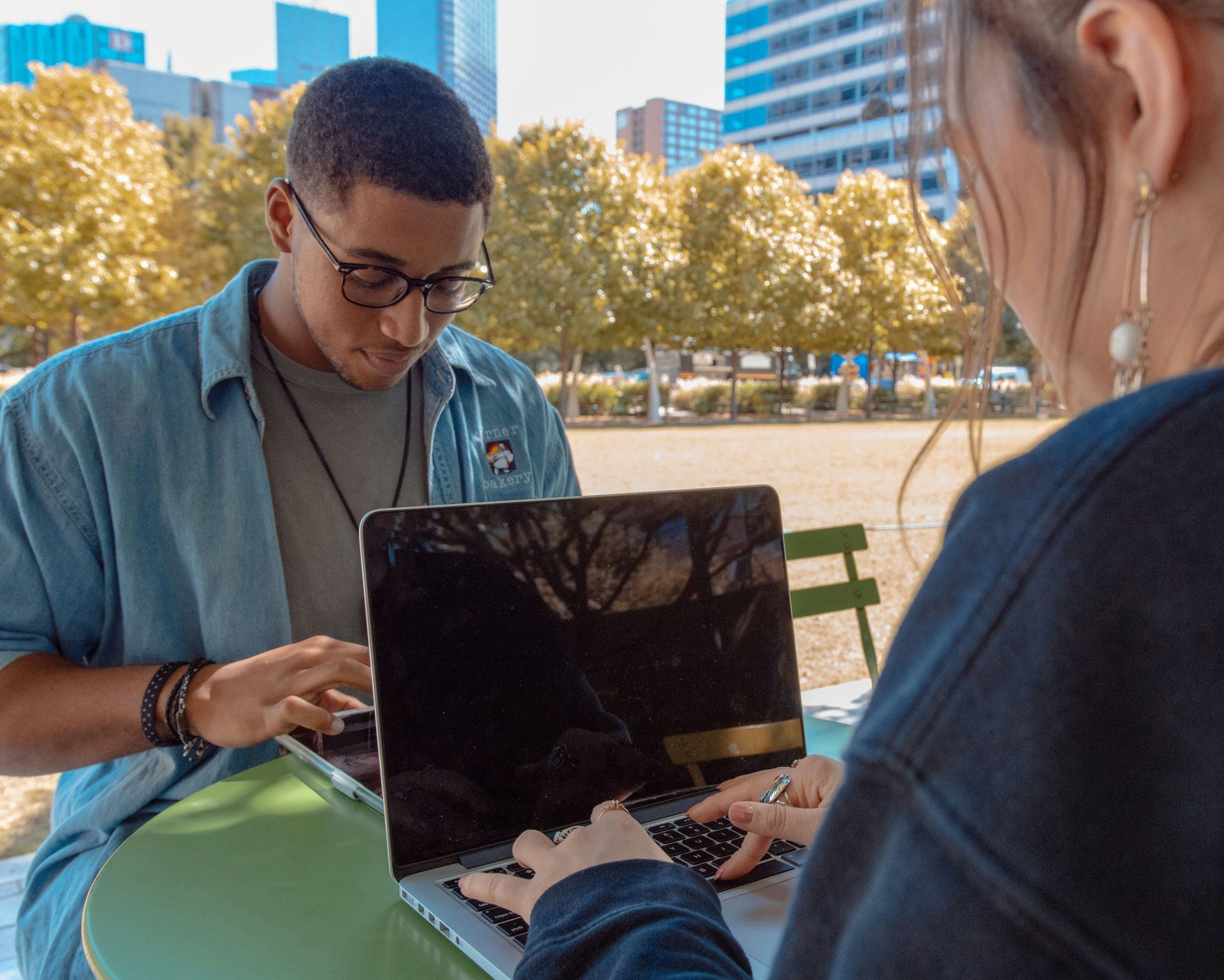 Two people work on laptops at an outdoor table; green table, trees, and buildings are in the background.