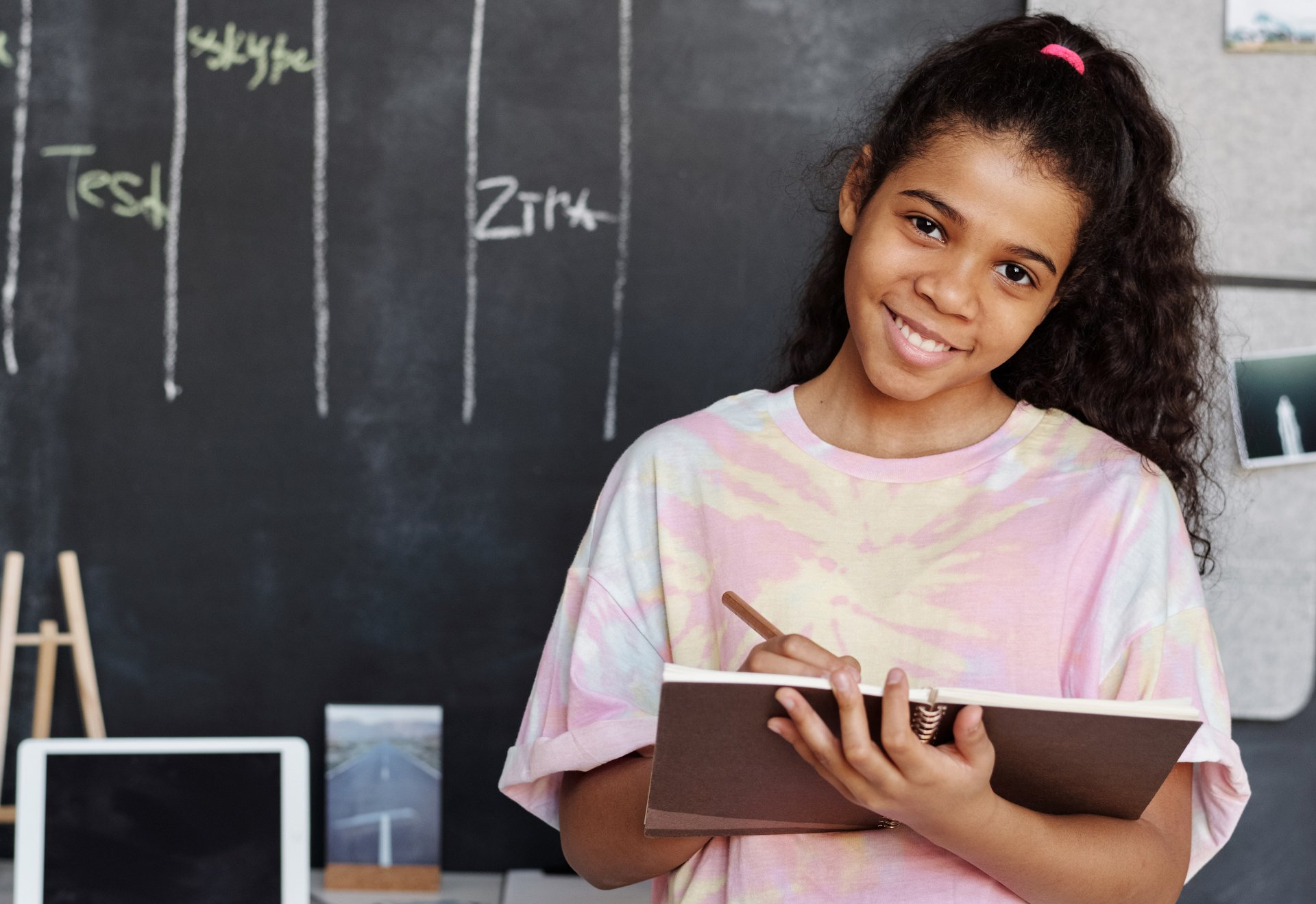 Smiling person writing in notebook in front of chalkboard with text.