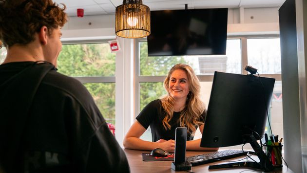 Een vrouw zit aan een bureau voor een computer en praat met een man.