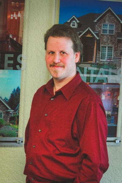 A man in a red shirt stands in front of a wall with pictures of houses on it