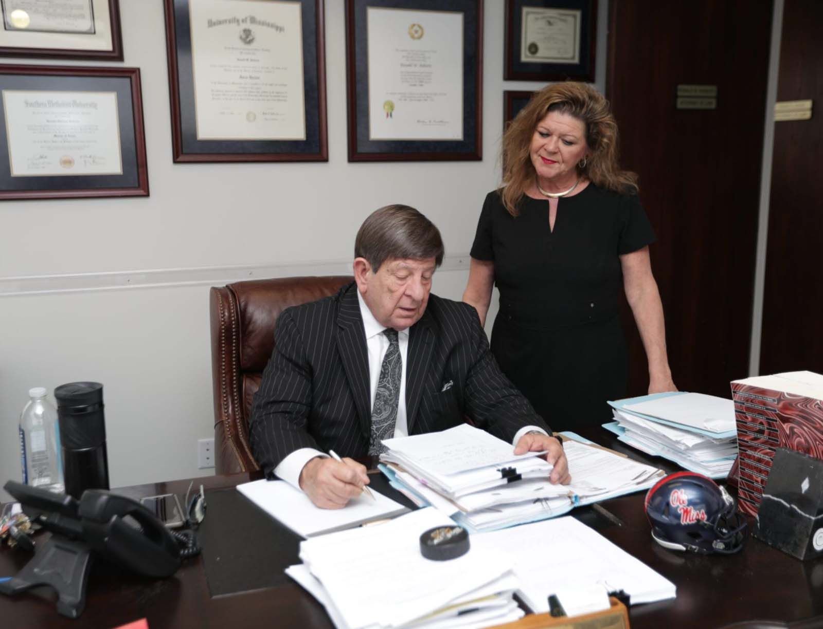 A man in a suit and tie is sitting at a desk with a woman standing behind him.