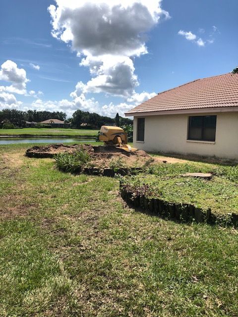 A yellow stump grinder sits on a mound of dirt next to a suburban house with a tiled roof and a nearby pond.