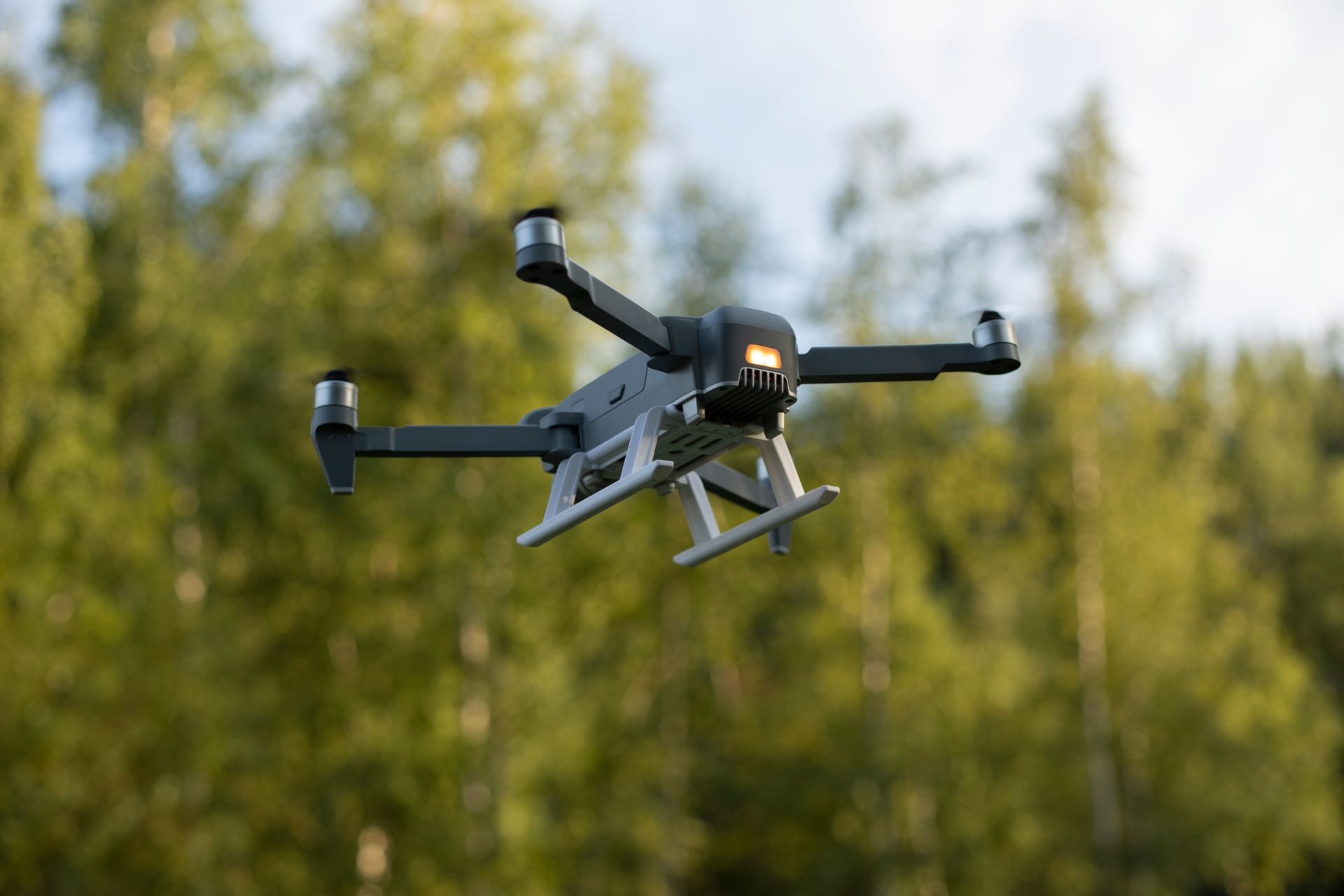 A dark gray drone with specialized white landing gear hovering in front of a blurred green forest.