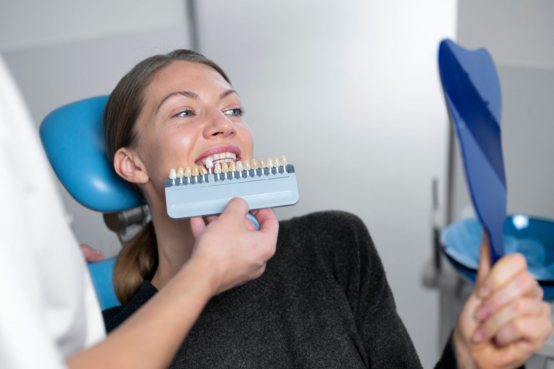 Woman in dentist chair comparing tooth shade to a color chart, holding a mirror.
