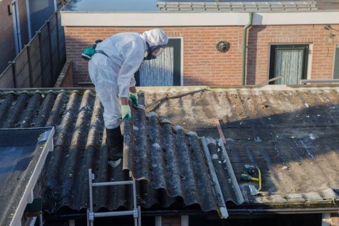 Worker in white protective suit repairing a corrugated roof beside a brick building.