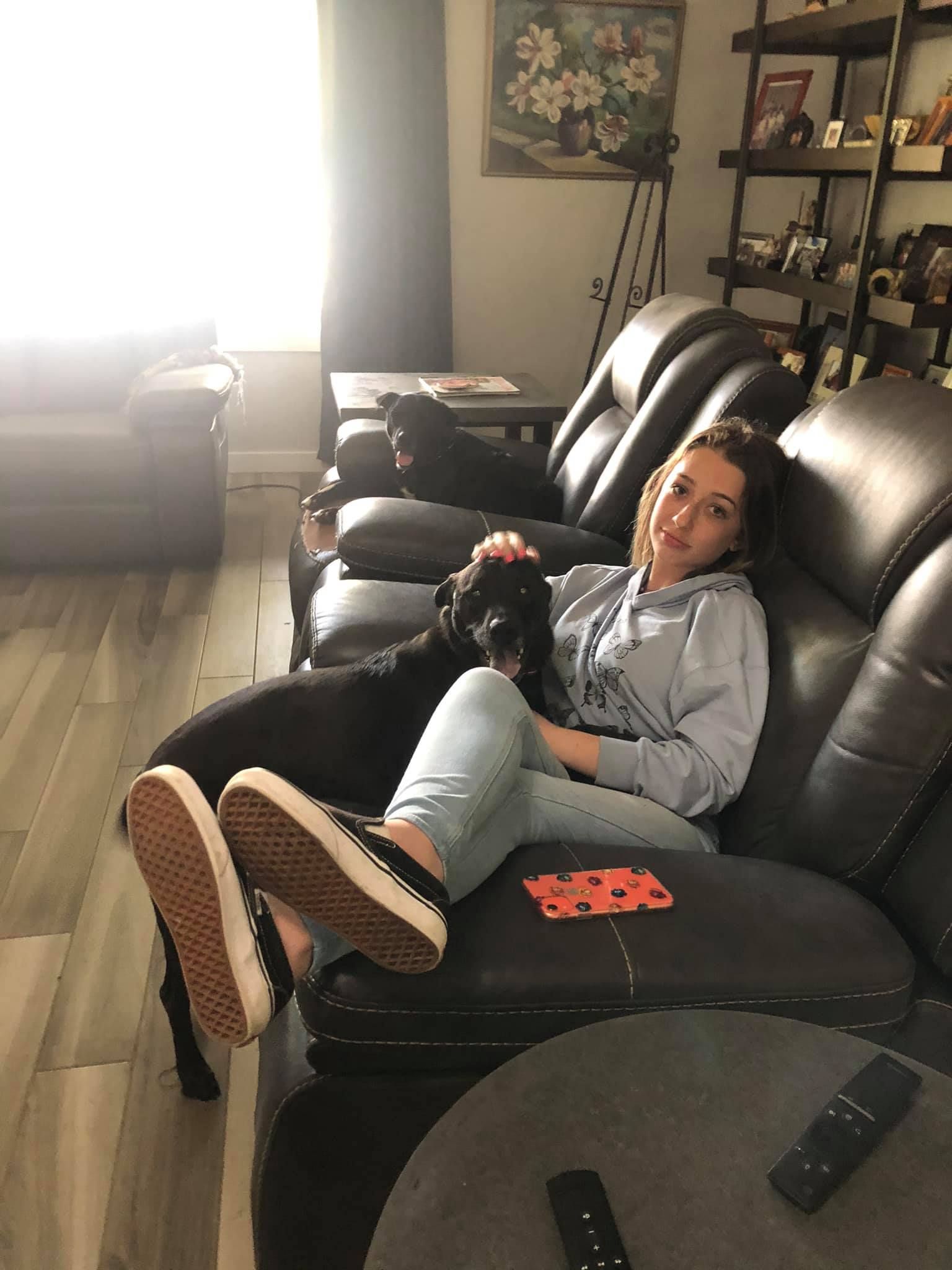 Woman on a black couch with two black dogs. She wears a light blue hoodie and jeans, with black sneakers. Inside.