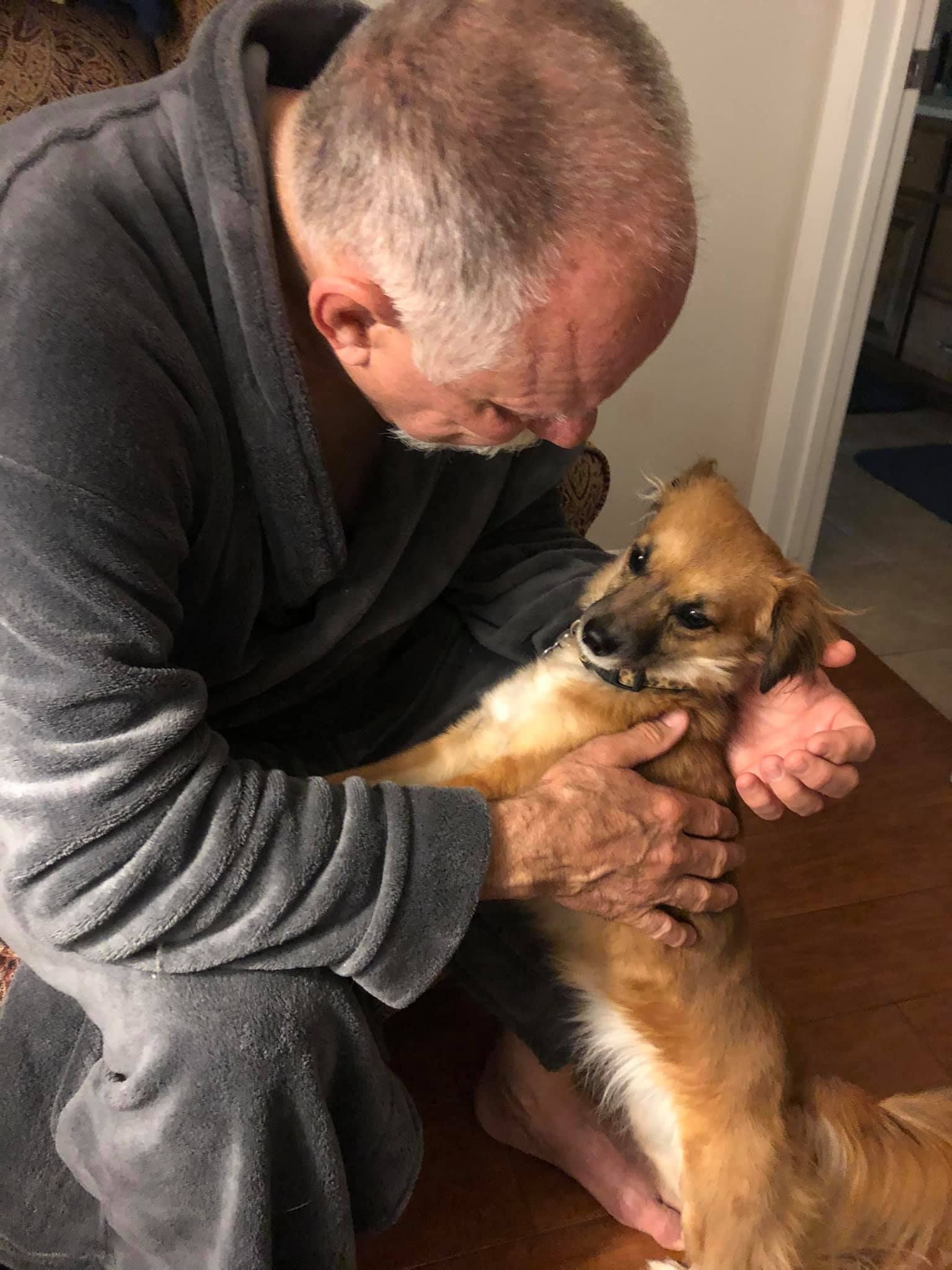 Man in gray robe holds a small, brown and tan dog, looking down with a gentle expression. Indoors.