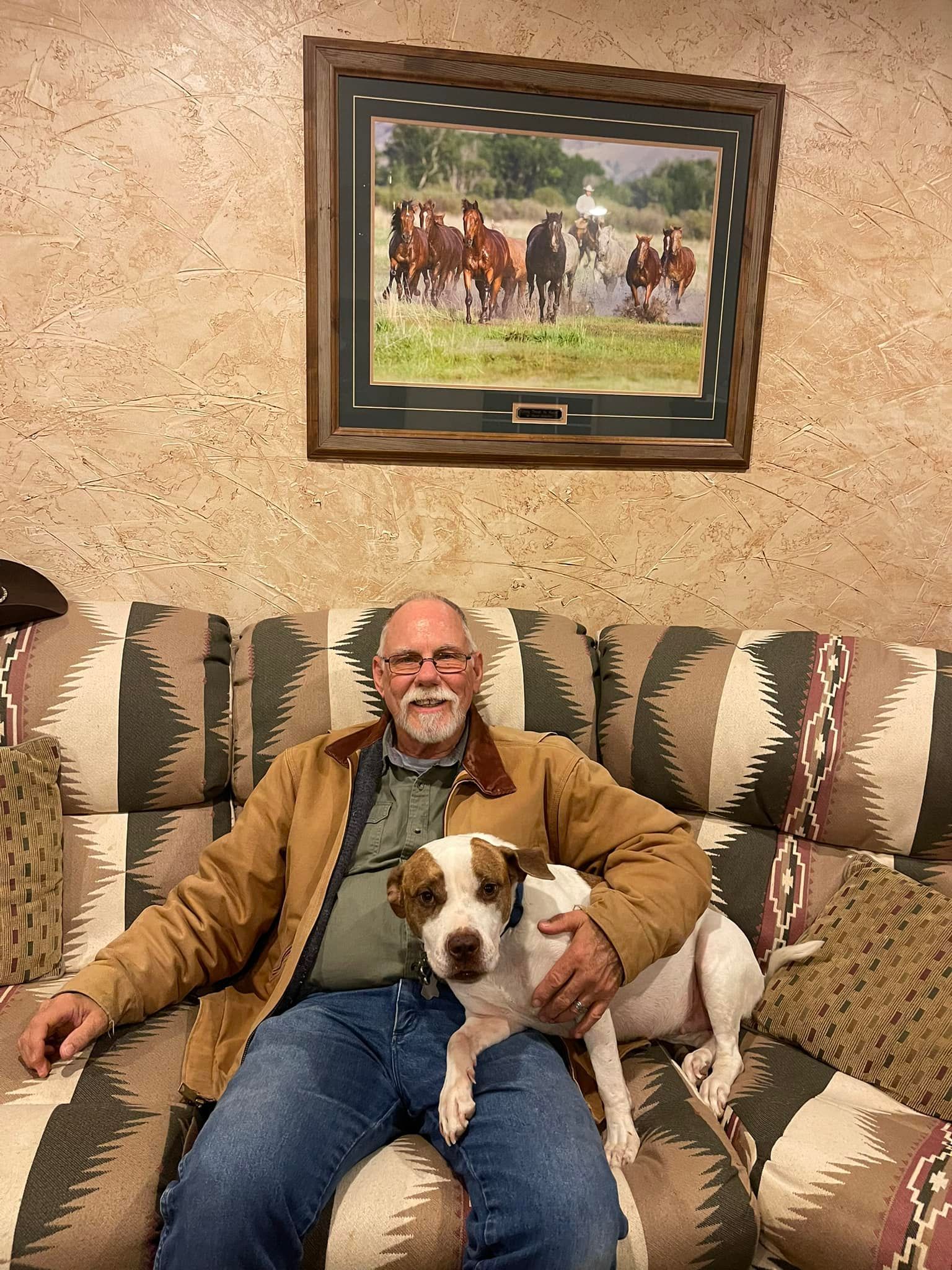 Man with dog on a southwestern patterned couch, picture of horses on wall. Man wearing brown jacket, jeans.