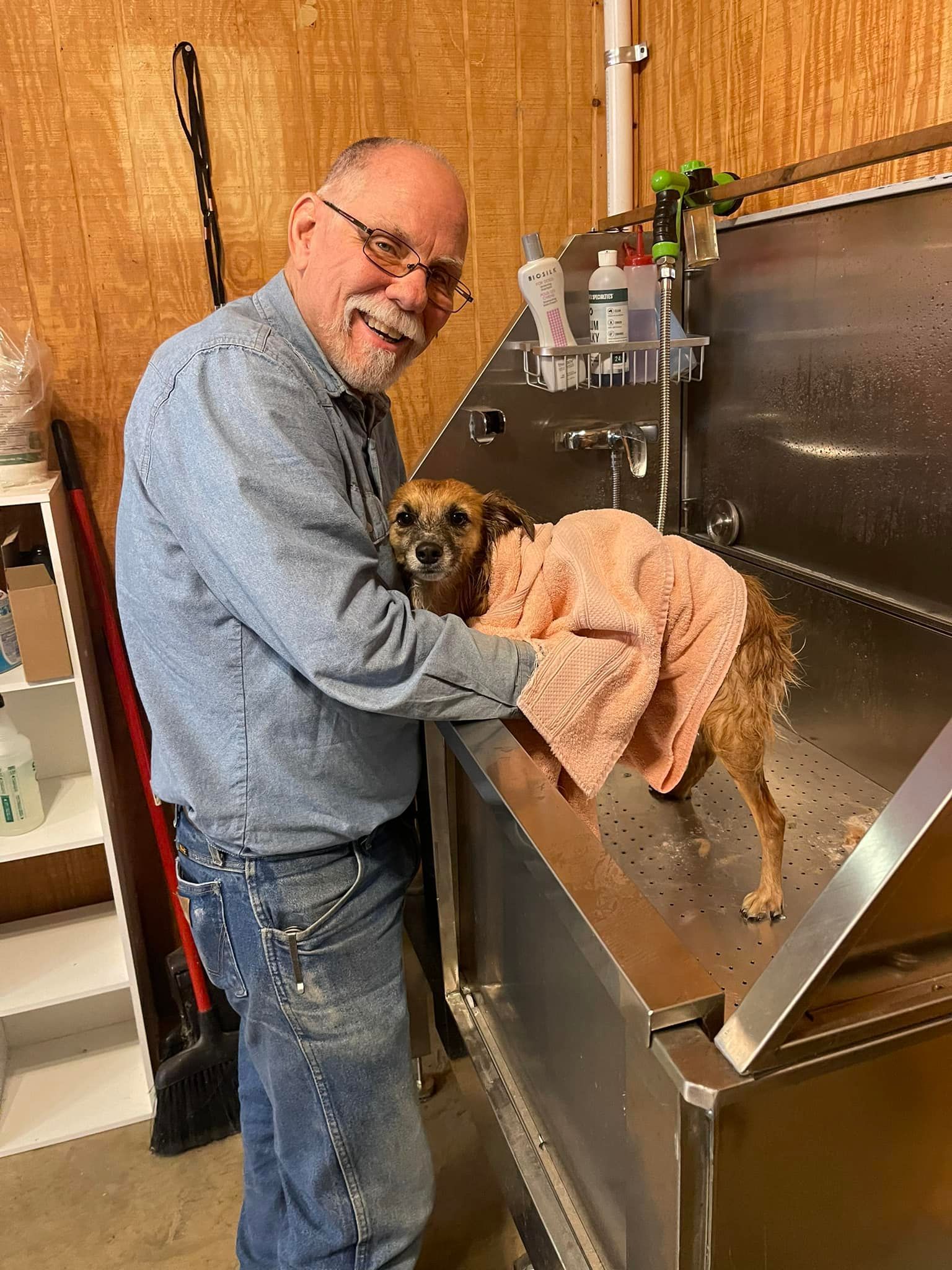 Man drying a small dog in a metal tub. The man wears glasses and a denim shirt. Brown dog wrapped in a pink towel.