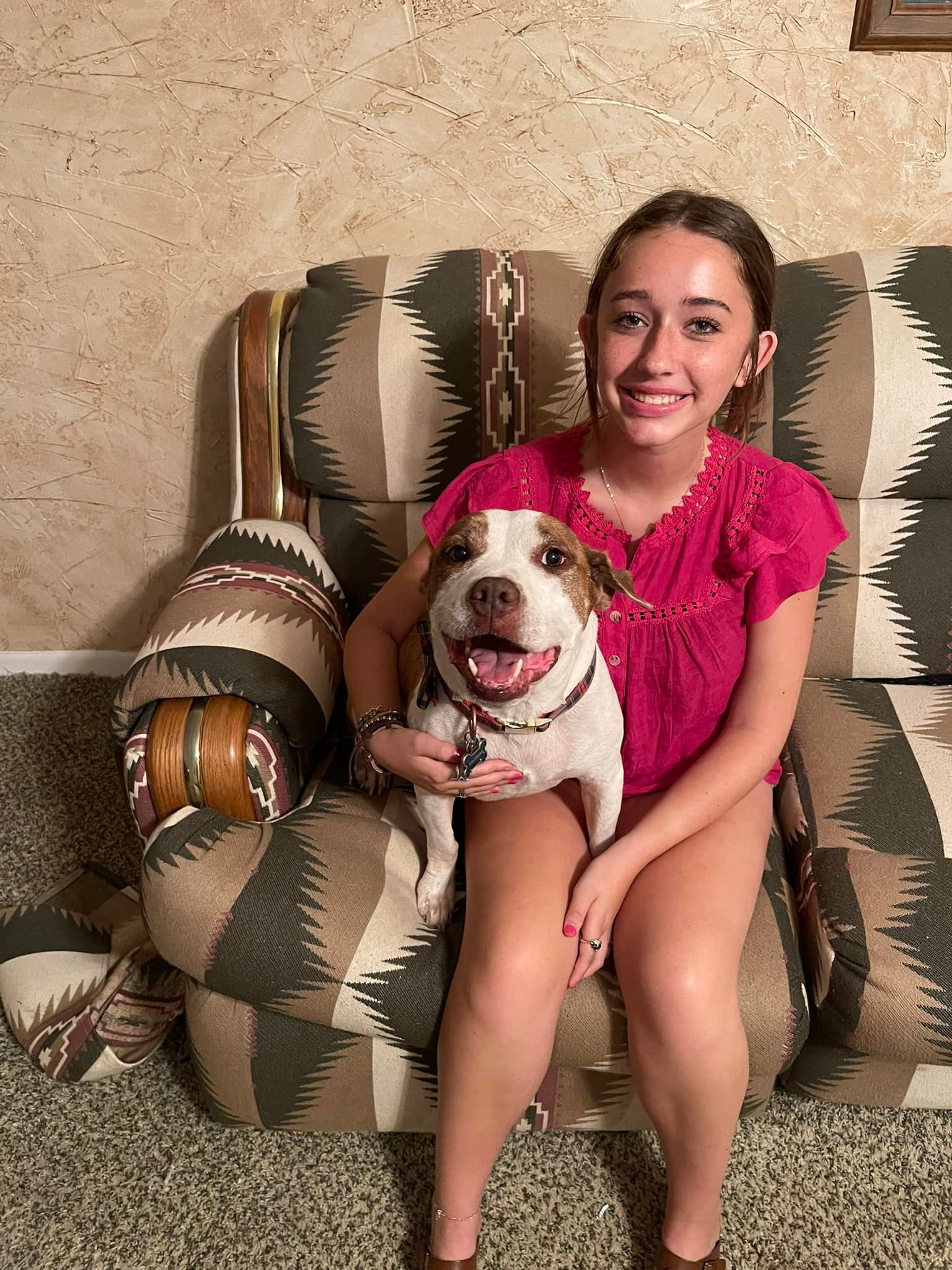 A young person smiles, holding a happy dog on a patterned chair. They are both inside.