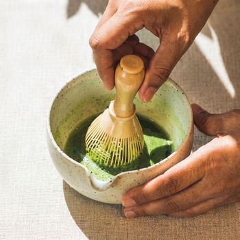 Hands whisking green matcha in a ceramic bowl with a bamboo whisk