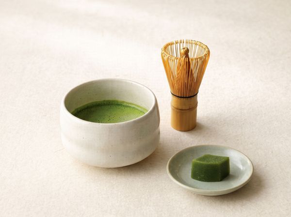 Matcha tea bowl, bamboo whisk, and a small green sweet on a plate.