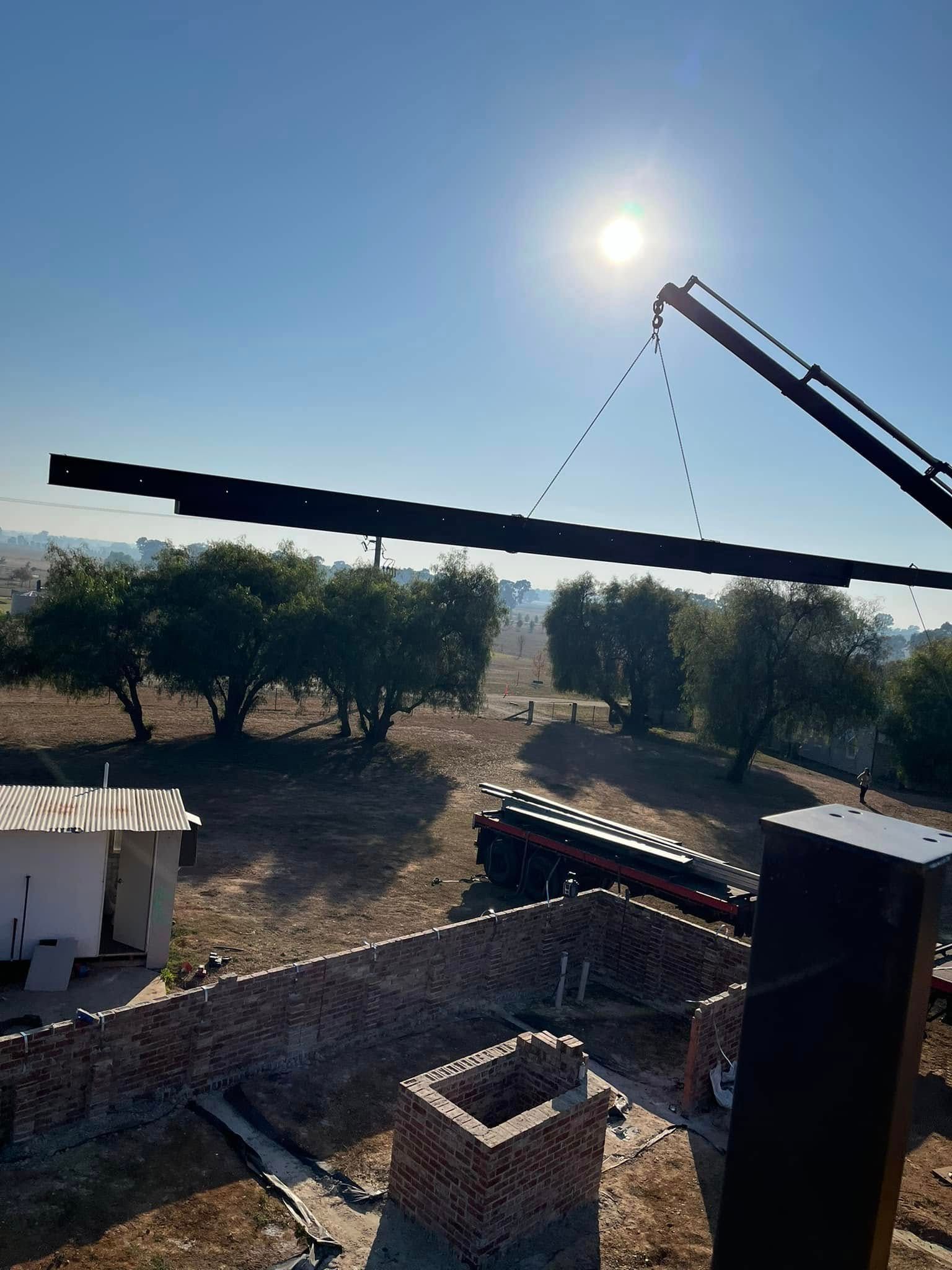 Construction site with crane lifting steel beam, sunny day. Brick walls and trees in the background.