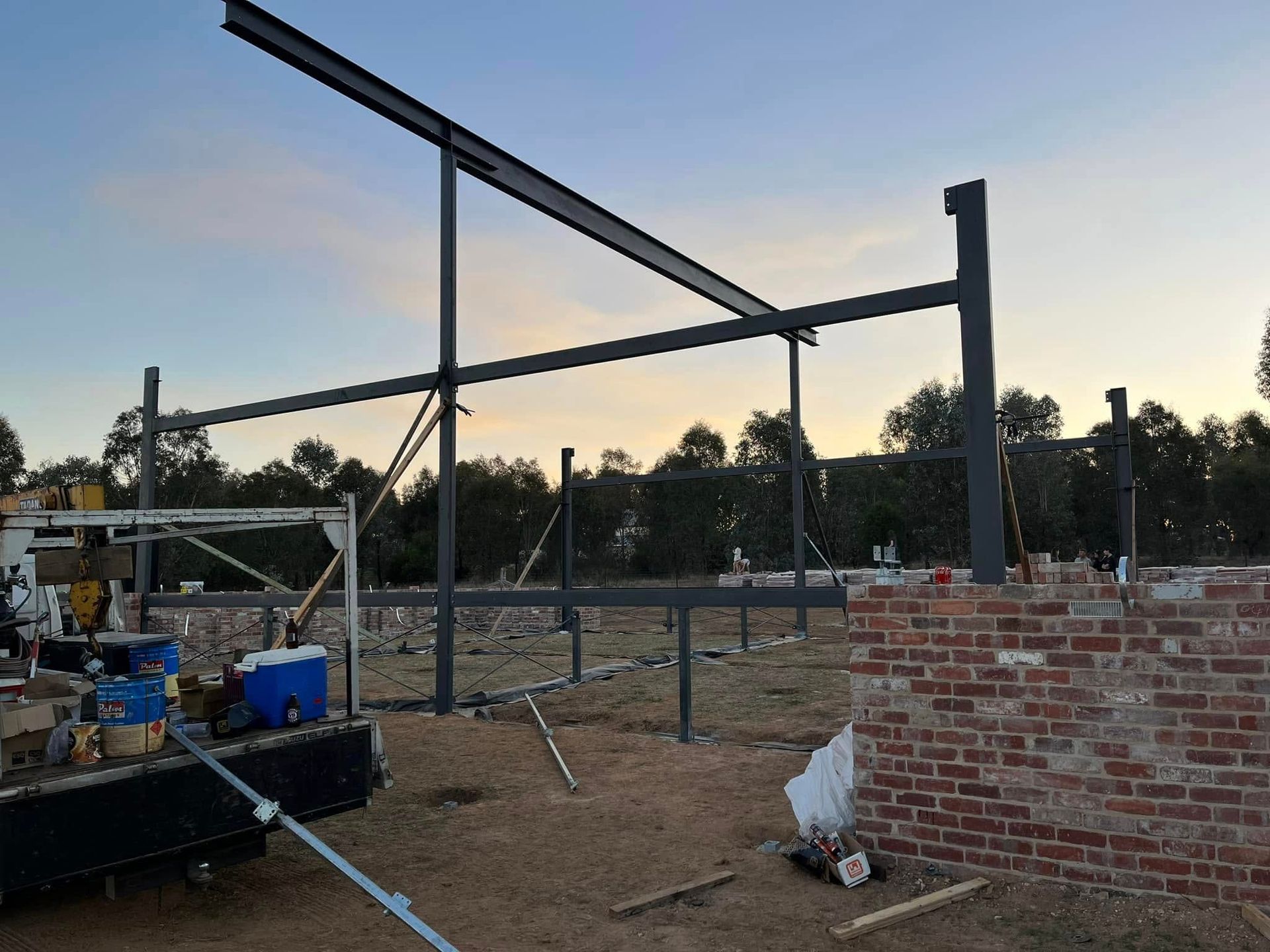 Steel frame of a building under construction, early evening.  Columns and beams against a light sky, partial brick wall visible.