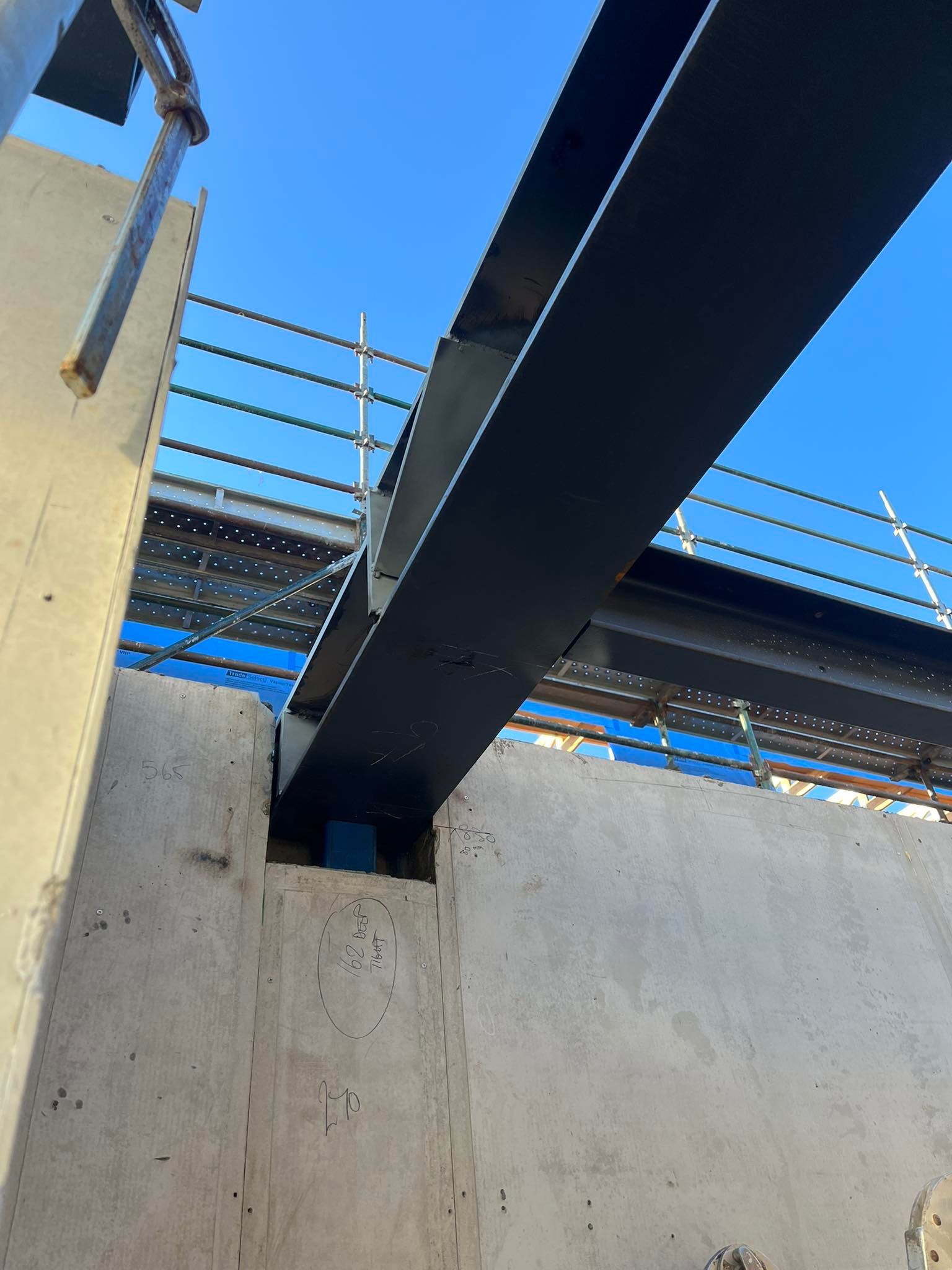 Construction site: Black steel beam spanning between concrete walls, scaffolding in background, blue sky.